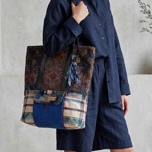 May include: A large tote bag featuring a dark patterned tapestry upper section, a plaid fabric lower section, and a solid blue central pocket with a button closure. The bag includes black leather-like shoulder straps and a blue tassel accessory.