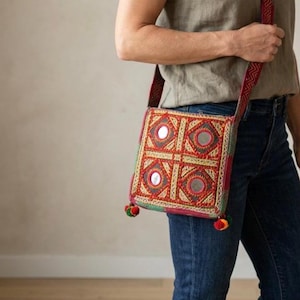 May include: A square, embroidered crossbody bag with a red, gold, and green geometric pattern. The bag features small, round mirrors and a red strap with a zigzag design. Small, colorful pom-poms adorn the bottom corners.