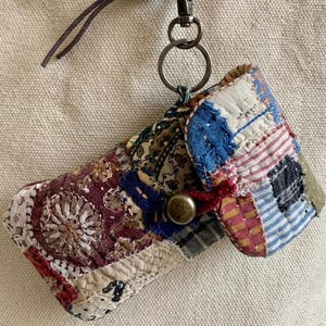 May include: A small rectangular pouch made of quilted patchwork fabric in various colours including blue, red, and cream. The pouch features a brass-toned button closure and is attached to a dark brown leather cord with a metal lobster clasp and ring.