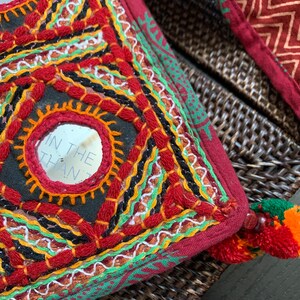 May include: Close-up of a decorative, embroidered pillow with a burgundy background. The pillow features geometric patterns with red, orange, black, and white stitching, and small mirrors. A tassel with orange and red accents is visible.