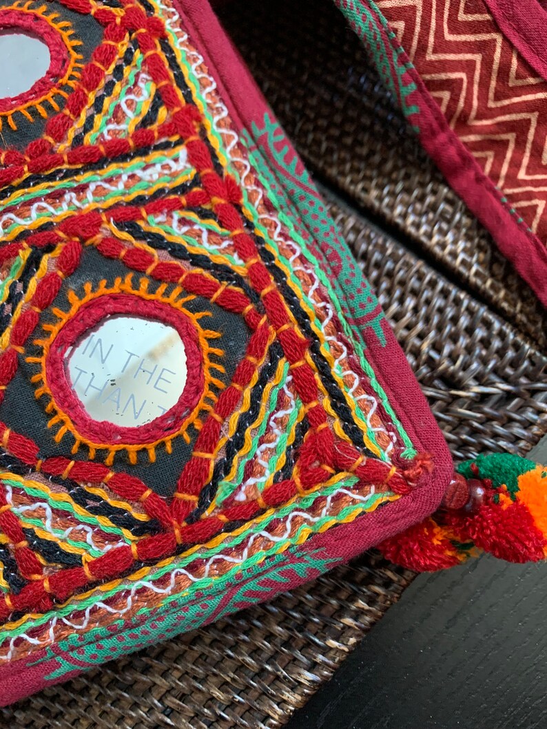 May include: Close-up of a decorative, embroidered pillow with a burgundy background. The pillow features geometric patterns with red, orange, black, and white stitching, and small mirrors. A tassel with orange and red accents is visible.