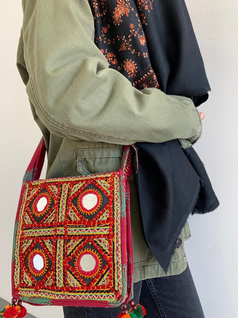 May include: A red and black embroidered shoulder bag with four mirrored squares. The bag has colorful stitching, a red strap, and decorative tassels. The bag is worn over a green jacket and black clothing.