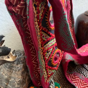 May include: A burgundy shoulder bag with intricate embroidery in orange, green, and yellow. The bag features a black base with mirror accents and a long strap with green patterns. Two small pom-poms hang from the bottom. The bag is a unique, handcrafted accessory.