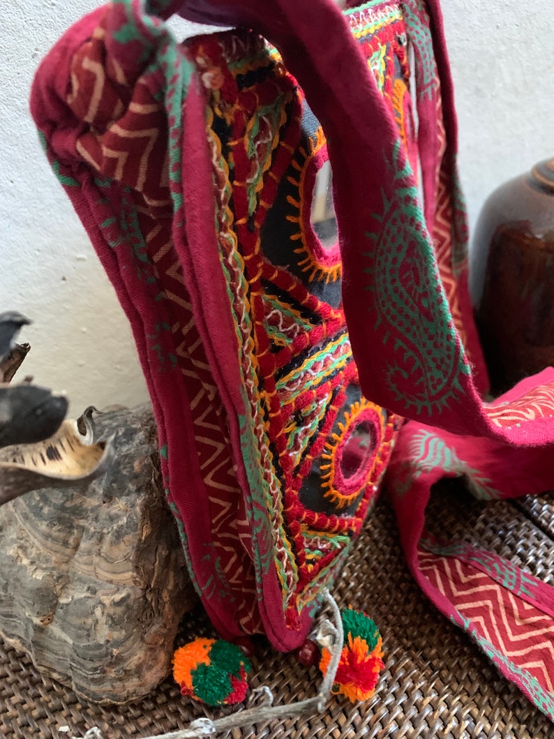 May include: A burgundy shoulder bag with intricate embroidery in orange, green, and yellow. The bag features a black base with mirror accents and a long strap with green patterns. Two small pom-poms hang from the bottom. The bag is a unique, handcrafted accessory.