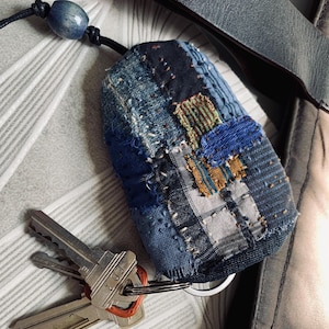 Può includere: Una piccola custodia per chiavi arrotondata realizzata in varie tonalità di tessuto denim blu, con cuciture a vista in stile sashiko e dettagli patchwork. La custodia è fissata a un cordino in pelle nera con una perla in legno blu e un anello.