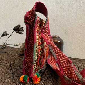 May include: A burgundy textile bag with a geometric pattern and colorful embroidery. The bag has a handle and is adorned with two pom-poms in orange and green. The design features a zig-zag pattern in a lighter shade of red.