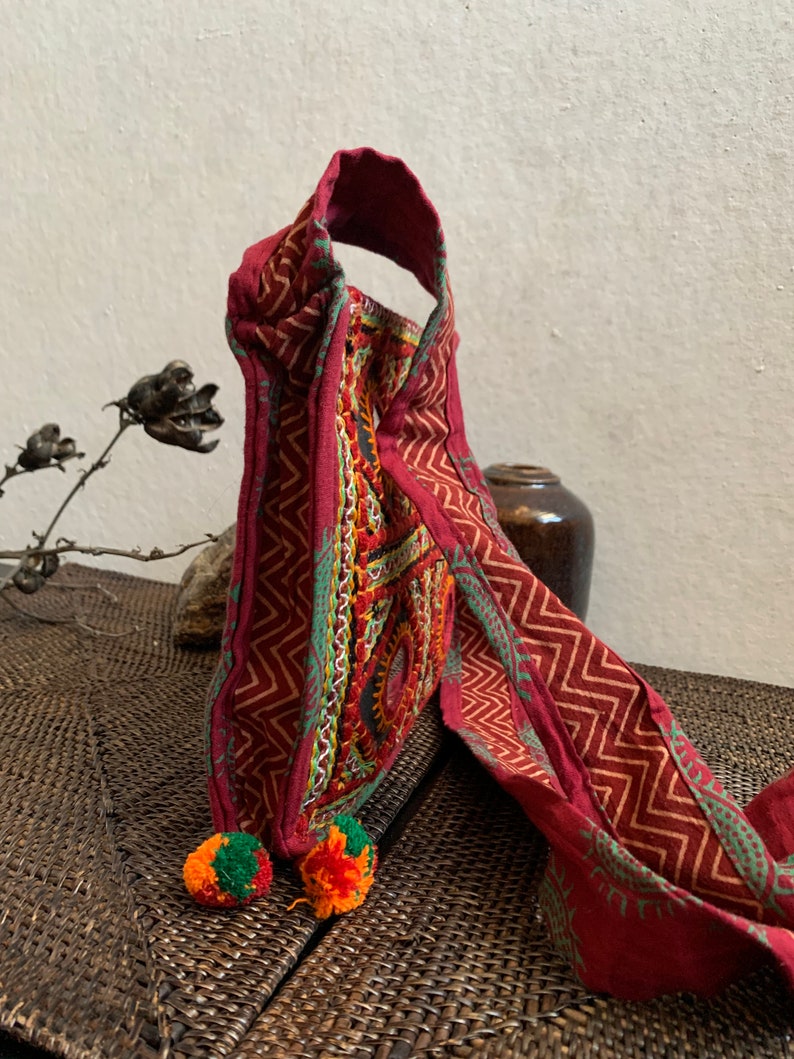May include: A burgundy textile bag with a geometric pattern and colorful embroidery. The bag has a handle and is adorned with two pom-poms in orange and green. The design features a zig-zag pattern in a lighter shade of red.
