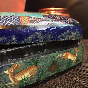 Peut inclure: Une boîte en bois peinte en bleu et vert avec un couvercle noir. La boîte est décorée d'un motif floral et de deux cerfs courant dans un champ. La boîte est posée sur une surface tissée.