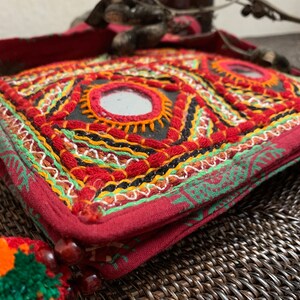 May include: A red, embroidered bag with geometric patterns and small mirrors. The bag features intricate stitching in red, green, yellow, and black. A colorful tassel with orange and green accents hangs from the side. The bag is a unique, handcrafted accessory.