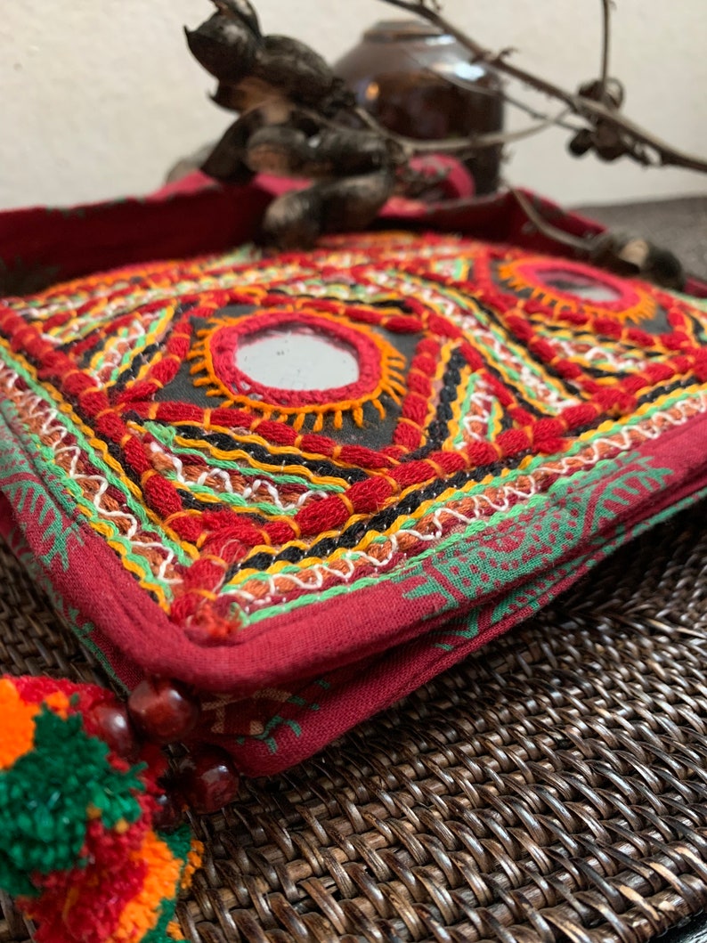 May include: A red, embroidered bag with geometric patterns and small mirrors. The bag features intricate stitching in red, green, yellow, and black. A colorful tassel with orange and green accents hangs from the side. The bag is a unique, handcrafted accessory.