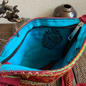 May include: Open view of a colorful, embroidered bag with a turquoise interior. The bag features a black zipper, intricate stitching in red, yellow, and green, and a paisley design on the inside. The bag is made of a red fabric with a patterned design.