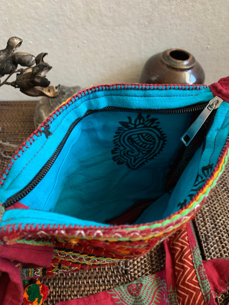 May include: Open view of a colorful, embroidered bag with a turquoise interior. The bag features a black zipper, intricate stitching in red, yellow, and green, and a paisley design on the inside. The bag is made of a red fabric with a patterned design.