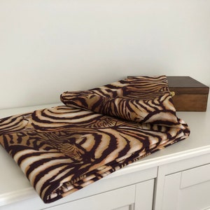 May include: A brown and tan zebra print fabric folded on a white surface. The fabric is folded in half and the edges are visible.