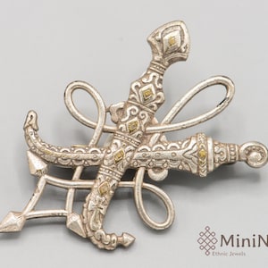 May include: Silver and gold tone brooch featuring two crossed daggers with intricate details. The daggers are intertwined with a decorative knot.