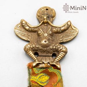 Puede incluir: Un adorno de metal dorado que representa una criatura mítica con alas y un cuerpo humanoide. El adorno está unido a un borla de tela colorida.