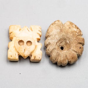 Puede incluir: Dos objetos de piedra tallada. Uno es un animal estilizado de color beige claro con dos agujeros circulares para los ojos. El otro es un objeto marrón en forma de flor con un agujero central. Ambos objetos tienen un aspecto envejecido.