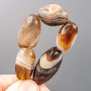 May include: A strand of six oval-shaped, polished agate beads. The beads display a range of colours, including brown, tan, and cream, with natural banding patterns. The beads are strung together with a thin, reddish-brown cord.
