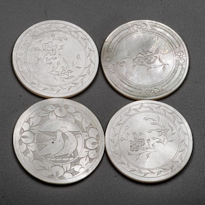 May include: Four round, mother-of-pearl coasters with engraved floral and bird designs. Each coaster features a unique pattern of flowers, leaves, and birds, creating an elegant and decorative set. The coasters are a light, iridescent color.