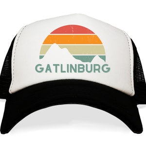 May include: A black and white trucker hat with a white front panel featuring a retro design of a mountain range and a sunset in red, orange, and yellow. The word "GATLINBURG" is printed below the mountains in green.