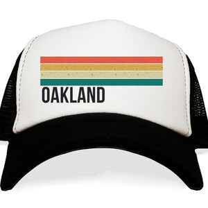 May include: A white and black trucker hat with a retro-style striped design in red, orange, yellow, and green. The text "OAKLAND" is printed in black below the stripes.