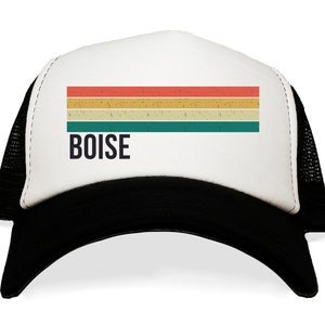 May include: A white trucker hat with a black mesh back. The front of the hat features a vintage-style striped design in red, yellow, and green colors. The word "BOISE" is printed in black below the stripes.