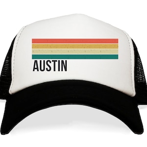 May include: A white and black trucker hat with a retro-style striped design in red, yellow, and green. The hat has the word "AUSTIN" printed in black on the front.