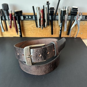 May include: A dark brown leather belt with a distressed, textured finish and a rectangular antique brass-toned metal buckle. The belt features a single leather loop and multiple punched holes for adjustment, coiled into a circular shape.