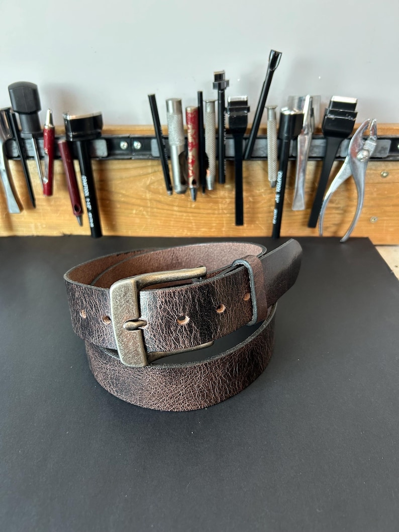 May include: A dark brown leather belt with a distressed, textured finish and a rectangular antique brass-toned metal buckle. The belt features a single leather loop and multiple punched holes for adjustment, coiled into a circular shape.
