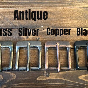 May include: Five metal belt buckles in different finishes: antique brass, silver, copper, and black. The buckles are arranged in a row from left to right.