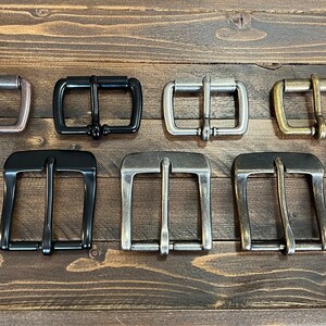 May include: A collection of nine metal belt buckles arranged in two rows. The top row features four smaller rectangular buckles in copper, black, silver, and brass. The bottom row contains five larger, wider buckles in copper, black, and silver tones.