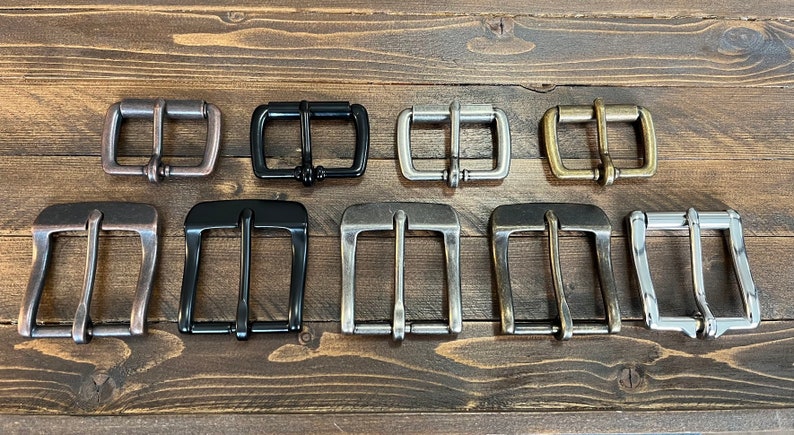 May include: A collection of nine metal belt buckles arranged in two rows. The top row features four smaller rectangular buckles in copper, black, silver, and brass. The bottom row contains five larger, wider buckles in copper, black, and silver tones.