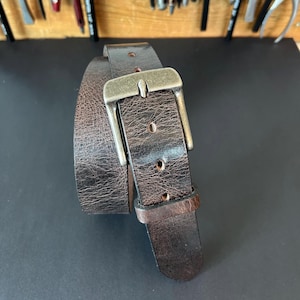 May include: A dark brown leather belt with a distressed, textured finish and a rectangular, antique-style metal buckle. The belt features a single leather keeper loop and multiple punched holes along the strap for adjustable sizing.