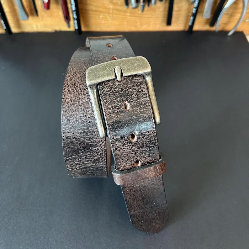 May include: A dark brown leather belt with a distressed, textured finish and a rectangular, antique-style metal buckle. The belt features a single leather keeper loop and multiple punched holes along the strap for adjustable sizing.