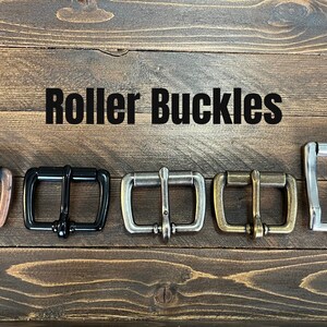May include: Five roller buckles in different finishes: copper, black, silver, brass, and nickel. The buckles are arranged in a row on a wooden surface. The text "Roller Buckles" is displayed above the buckles.