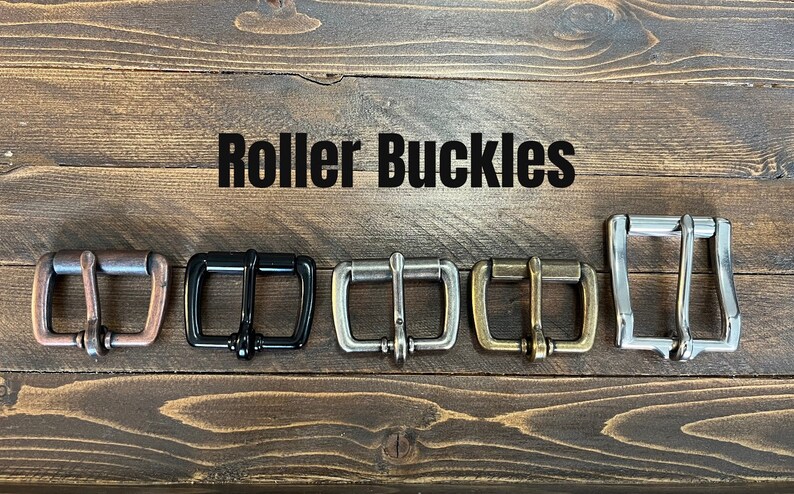 May include: Five roller buckles in different finishes: copper, black, silver, brass, and nickel. The buckles are arranged in a row on a wooden surface. The text "Roller Buckles" is displayed above the buckles.