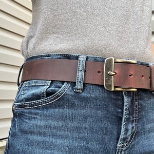 May include: A brown leather belt with a brass buckle. The belt is worn over a pair of blue jeans.