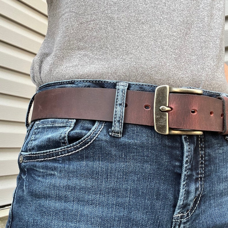 May include: A brown leather belt with a brass buckle. The belt is worn over a pair of blue jeans.