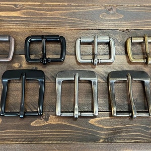 May include: A set of ten metal belt buckles in various finishes, including antique brass, silver, black, and nickel. The buckles are all the same size and shape, with a rectangular frame and a double prong closure.