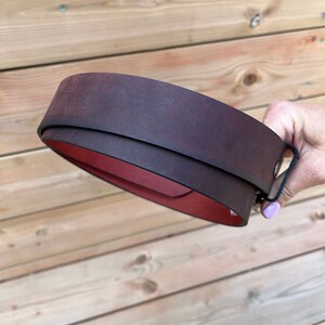 May include: A brown leather belt with a red lining. The belt is rolled up and held in a hand.