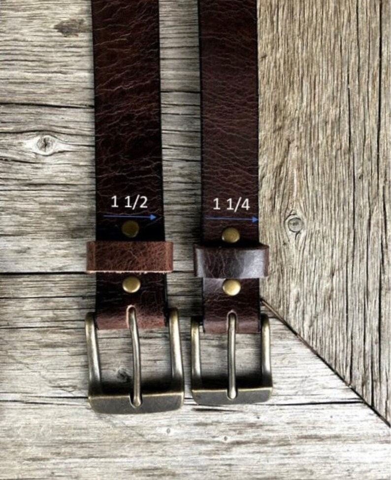 May include: Two dark brown textured leather belts with antique brass-toned metal buckles and rivets. The left belt features a width measurement of 1 1/2 inches, while the right belt features a width measurement of 1 1/4 inches as indicated.