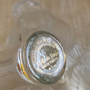 May include: A clear glass bottle with a gold label that reads "Parfums Nina Ricci - Paris - Vaporisateur Spray - L'Air du Temps - Eau de Toilette - 82% vol. - Made in France - Cont. 30 ml."