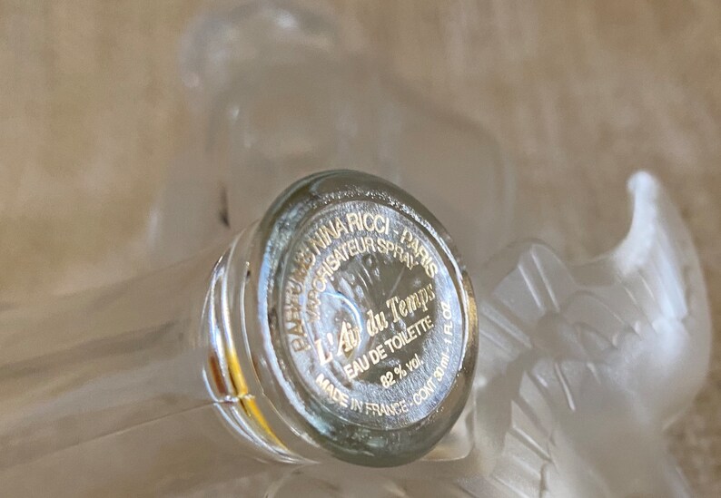 May include: A clear glass bottle with a gold label that reads "Parfums Nina Ricci - Paris - Vaporisateur Spray - L'Air du Temps - Eau de Toilette - 82% vol. - Made in France - Cont. 30 ml."