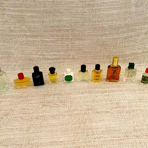 Pode incluir: Uma coleção de frascos de perfume em miniatura de várias formas e cores. Os frascos estão dispostos em fila, com vidro transparente, âmbar e preto. Alguns têm tampas prateadas, vermelhas e verdes. Os frascos estão sobre um tecido de cor neutra.