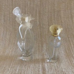 May include: Two clear glass perfume bottles with decorative stoppers. The bottle on the left has a white angel-shaped stopper. The bottle on the right has a gold-colored stopper with a floral design.