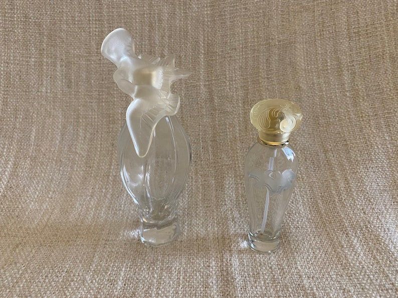 May include: Two clear glass perfume bottles with decorative stoppers. The bottle on the left has a white angel-shaped stopper. The bottle on the right has a gold-colored stopper with a floral design.