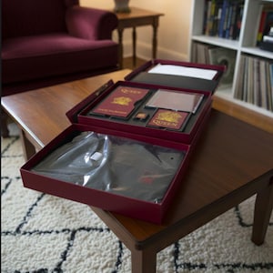 Puede incluir: Un set de caja granate sobre una mesa de madera, que contiene una camiseta negra, un CD y otros recuerdos. La caja tiene la palabra "Queen" impresa. El set está abierto, revelando el contenido. El fondo incluye un sillón granate y una estantería.