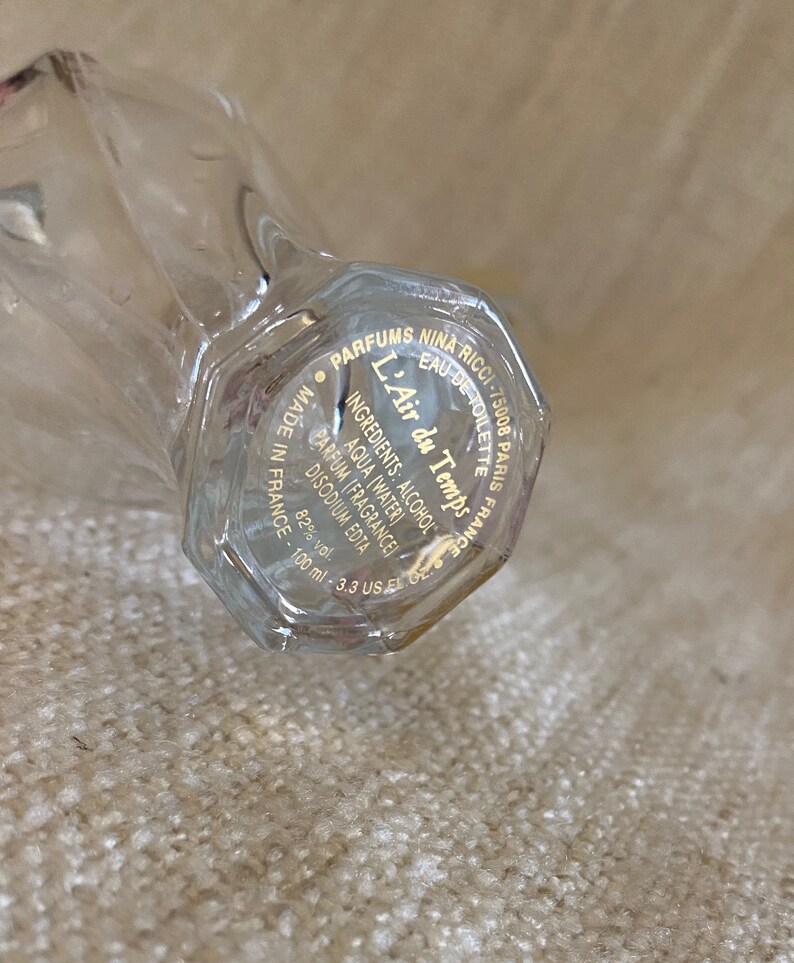 May include: A clear glass bottle with a gold label that reads "Parfums Nina Ricci - 75008 Paris France - Eau de Toilette - L'Air du Temps - Ingredients: Alcohol, Aqua (Water), Parfum (Fragrance), Disodium EDTA - 82% vol. - 100 ml - 3.3 US fl oz - Made in France"
