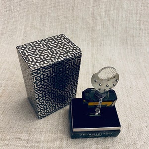 Puede incluir: Una caja negra y dorada con un patr&oacute;n geom&eacute;trico se encuentra junto a una peque&ntilde;a botella de perfume. La botella tiene una tapa de vidrio transparente con un tap&oacute;n plateado y una cadena plateada que cuelga de ella. La botella est&aacute; etiquetada como "Uninhibited Perfume".