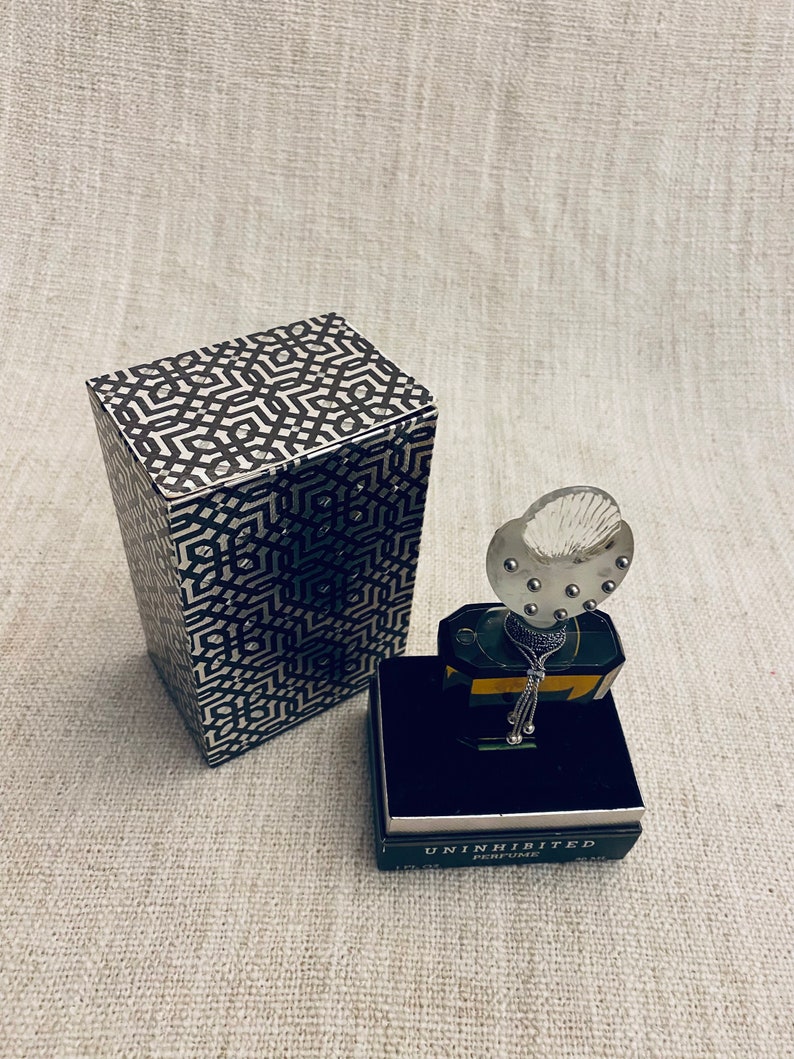 Puede incluir: Una caja negra y dorada con un patr&oacute;n geom&eacute;trico se encuentra junto a una peque&ntilde;a botella de perfume. La botella tiene una tapa de vidrio transparente con un tap&oacute;n plateado y una cadena plateada que cuelga de ella. La botella est&aacute; etiquetada como "Uninhibited Perfume".