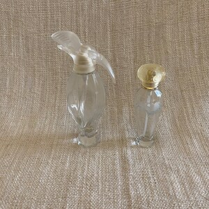 May include: Two clear glass perfume bottles with different shaped stoppers. The larger bottle has a clear, sculpted stopper shaped like a bird. The smaller bottle has a gold-colored, round stopper.